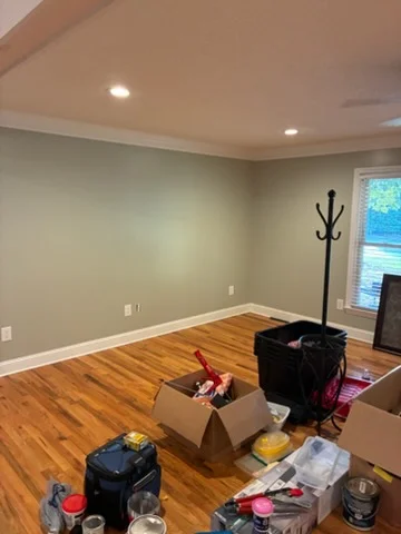 Sage green painted interior wall with white crown molding, white baseboards, and hardwood floor near paint cans and tools