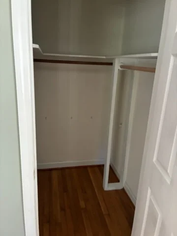 Walk-in closet with smooth white painted walls, white trim and shelf, dark wood hanging rod, and hardwood floor